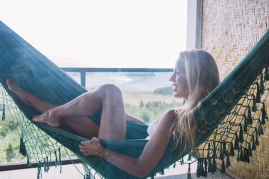 Güneşli bir günde bahçe girişi olan açık terasta mayo giymiş, hamakta dinlenen ve gözlerini başka yöne çeviren büyüleyici, bronz tenli kadının yan görüntüsü.