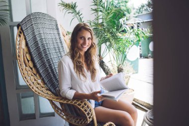 Hasır sandalyede oturan çekici genç bir kadın. Terasta ilginç bir kitabın keyfini çıkarırken gülümseyen ve kameraya bakan.