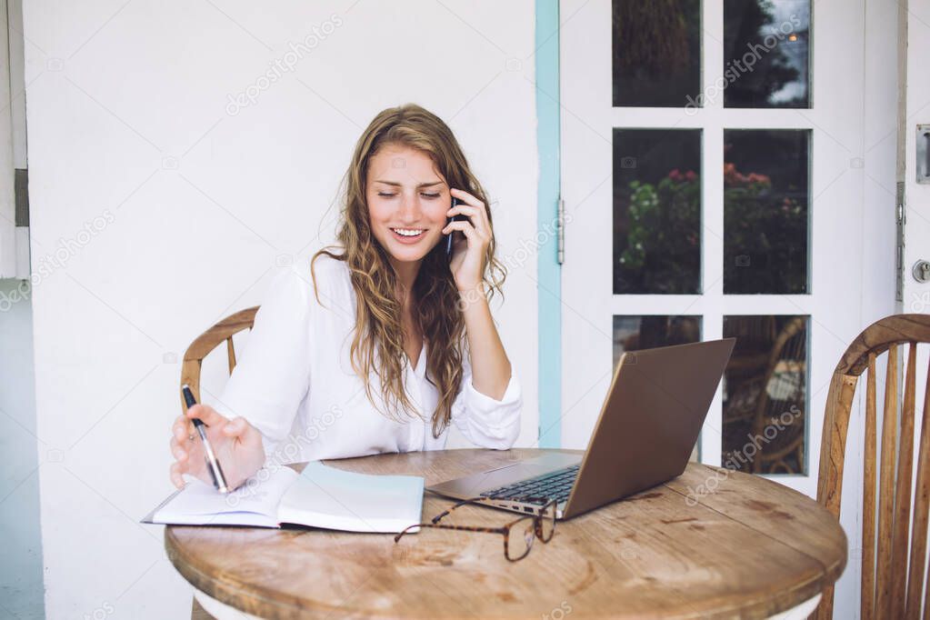 Cheerful attractive woman in white using notebook and talking on phone while sitting with laptop at rounded table in terrace 