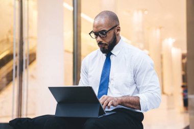 Resmi giysiler içinde düşünceli ve ciddi bir siyah işçi pencerenin yanında otururken tablet kullanan meslektaşlarına mesaj yazıyor. 