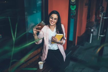 Ders kitabı yazan neşeli kadın şair molada cep telefonu ön kamerasını kullanarak selfie çekerken mutlu Gürcü kadın kaldırımda boş zamanlarında video çekiyor.
