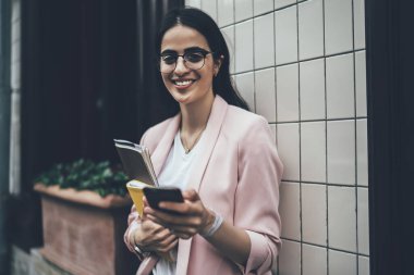 Başarılı İspanyol kullanıcının yarım boy portresi eğitim ekipmanları ve modern mobil teknoloji, optik gözlüklü zengin hippi kız boş zamanlarında kameraya gülümsüyor.