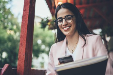 Başarılı İspanyol kullanıcının yarım boy portresi eğitim ekipmanları ve modern mobil teknoloji, optik gözlüklü zengin hippi kız boş zamanlarında kameraya gülümsüyor.