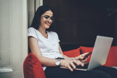 Laptop ve akıllı telefon teknolojisi arasında medya dosyalarını paylaşmak için bluetooth 'a bağlanan mutlu İspanyol kadın, serbest iş sırasında mobil tebligatı kontrol eden gözlüklerde başarılı bir milenyum