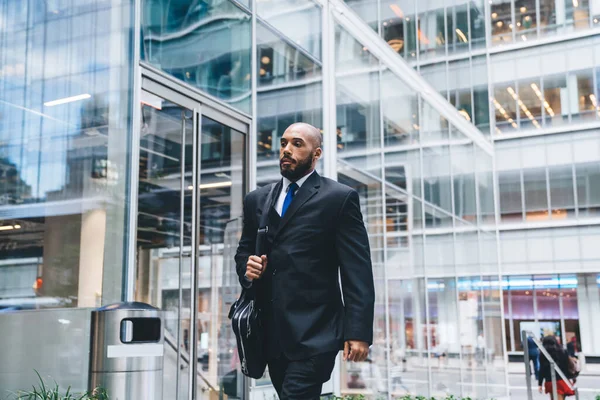 Confident bald bearded black businessman in suit holding black leather briefcase while walking in commercial building on background of glass windows