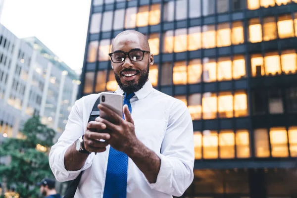 From below successful African American bald bearded employee with glasses messaging on white smartphone while standing in front of modern office building