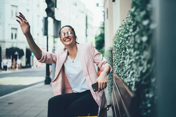 Cheerful Hispanic woman in classic eyewear for vision correction happy with unexpected live meeting in city, happy female 20s holding cellphone gadget and waving while waiting friend at bench