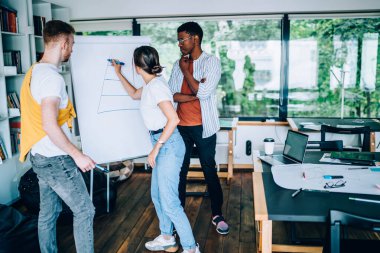 Gündelik kıyafet grafiği çizen genç bir kadın, çok ırklı meslektaşlarının yanında dikilip modern ofis projesinde çalışıyor.