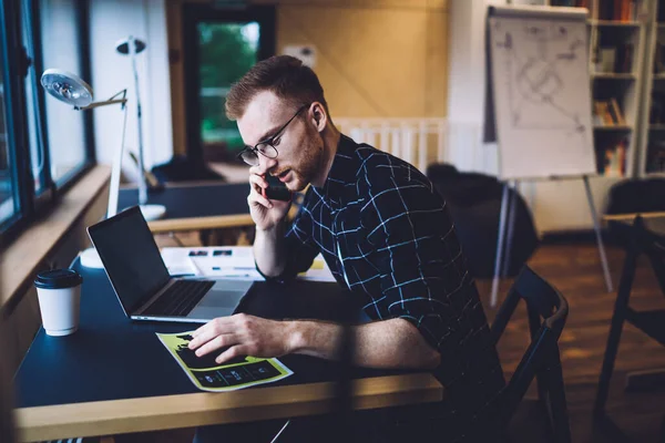 Caucasian male programmer sitting at table desktop and using mobility connection for making cellphone communication and discussing database details, smart casual copywriter phoning via gadget