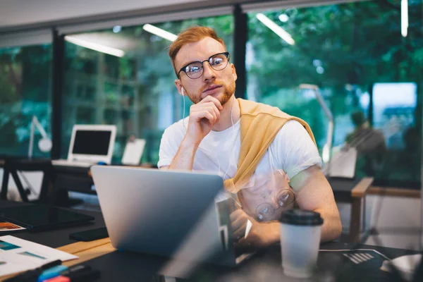 年轻聪明的 头戴眼镜 头戴红头发的男性 穿着休闲的白色T恤 手牵着下巴 坐在笔记本电脑旁 边喝咖啡边浏览 — 图库照片