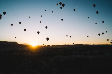 Sıcak hava balonlarının hava manzaralı siluetleri şafak vakti karanlık dağların üzerinde uçuyor ve ufukta güneş doğuyor açık gökyüzü var.
