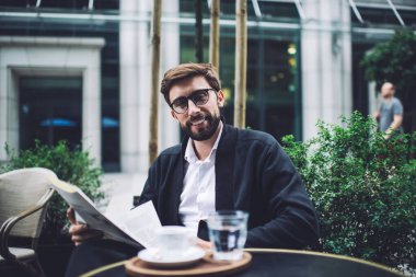 Neşeli sakallı, gözlüklü iyi giyimli, kameraya bakıp gülümseyen bir erkek sokak kafesinde gazete ve kahveyle oturuyor.