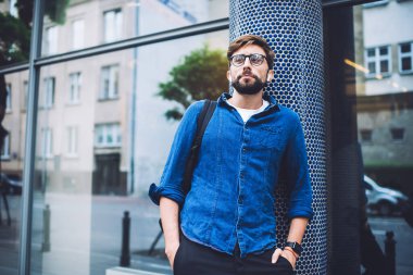 Sakallı, gözlüklü ve sırtında sırt çantası olan yaz günü sokakta aktiviteleri izleyen sıradan bir genç.