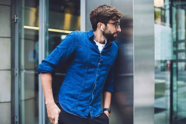 Gözlüklü ve kulaklıklı sakallı genç adam yazın sokakta dikilirken insanları köşeden izliyor.