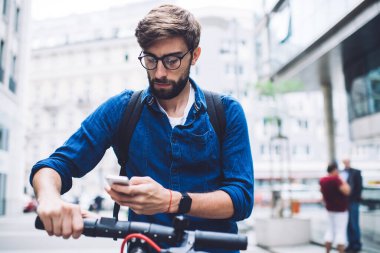 Kot gömlek ve kol saati giymiş, Scooter 'ın üzerinde dikilirken cep telefonuyla sohbet eden konsantre sakallı, genç bir erkek öğrenci.