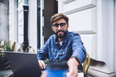 Kot ceketli ve gözlüklü neşeli bir erkek, güneşli bir günde açık netbook ile otururken isimsiz bir garsondan plastik bir fincan kahve alıyor.
