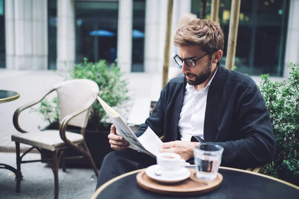 在街上的咖啡店里 身穿眼镜 留着优雅胡子 衣冠楚楚的男人一边认真地看报纸 一边喝着一杯热饮 — 图库照片