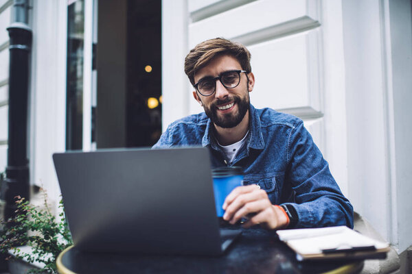 Satisfied young hipster male in casual wear and eyeglasses with laptop and clipboard enjoying takeaway coffee in cafe and looking at camera