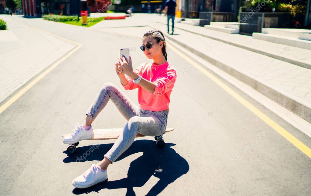 Millennial skater dressed in fashionable streetwear clicking content selfie during leisure in city, hipster girl sitting at longboard and creating influence vlog for sharing to social networks