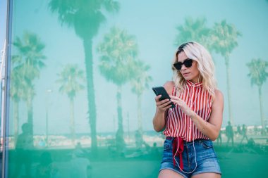 Moda giyinmiş, şık güneş gözlüklü yeniyetme kız internet mağazasından modern akıllı telefona geçmek için medya uygulamasını seçiyor. Elektronik kulaklıklı bin yıllık kadın online müzik podcast dinliyor.