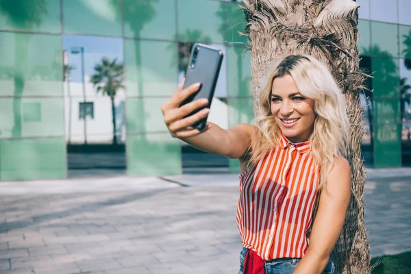 Happy Caucasian influence blogger smiling at mobile camera while shooting video vlog enjoying leisure near palm tree, cheerful millennial with blonde hair using cellphone app for selfie photographing