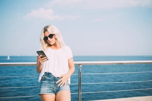 海辺の桟橋で夏の遊歩道のための時間の間にモビリティ通知を検索トレンディーなサングラスのミレニアル世代の女性 スマートフォンアプリ経由でメッセージングのために4Gに接続白人ヒップスター女の子 — ストック写真
