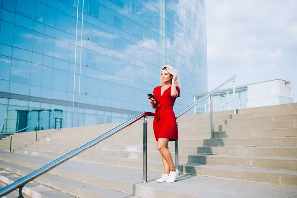 身穿时髦红色太阳衣的年轻女子站在城市的楼梯上 思索着网络共享的内容 千禧年嬉皮士女孩手握现代蜂窝式小玩艺儿 为流动休闲 — 图库照片