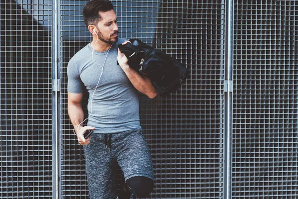 自信的高加索肌肉男运动员拿着时髦的背包 带着运动用具站在城市里 英俊而严肃的运动员在智能手机上听音乐 — 图库照片