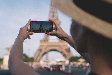 Güneşli bir günde Paris caddesinde güneşli bir günde Eiffel Kulesi 'nin dışını çekmek için akıllı telefon kullanan şapkalı isimsiz bir kadın.