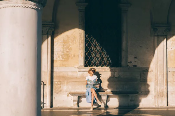 帽子とサングラスの幸せな若い女性が先を見て イタリアの日当たりの良いヴェネツィアのベンチで観光中に座っている間地図を保持 — ストック写真