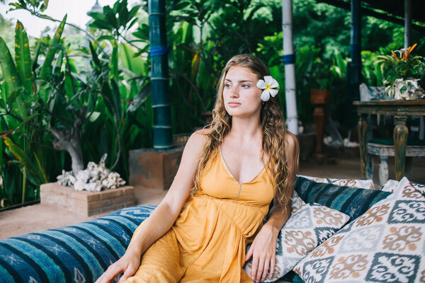 Меланхоличная взрослая блондинка с орхидеей в волосах в оранжевом платье смотрит в сторону и думает, сидя и опираясь на подушки на диване в тропическом саду на курорте Бали в солнечный день