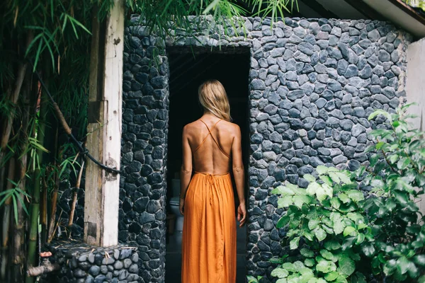 stock image Back view of gorgeous young woman in stylish orange dress standing on threshold of outdoor bathroom with pebble stone wall on Bali