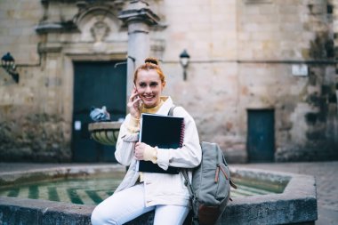 Sırt çantası ve defterleri olan mutlu genç bir kadın Barcelona 'nın antik meydanında çeşmenin yanında otururken gülümseyip cevap veriyor.