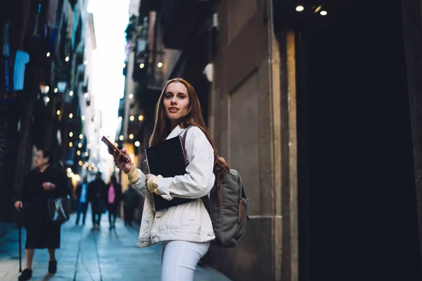 都会の通りに立っている間にスマートフォンを使用して暖かいジャケットでバックパックやコピー本とかなり笑顔穏やかな若い女性の側面図 — ストック写真