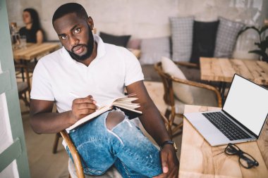 Eğitim kitaplarıyla çalışırken kameraya bakan koyu tenli erkek yazarın portresi boş dizüstü bilgisayarla masanın yanında oturuyor, web siteniz için kopyalama alanı olan modern model bilgisayar