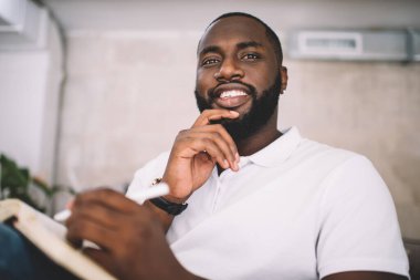 20 'li yaşlarda, neşeli, esmer bir adamın portresi. Not defterine yazı yazan, gülümseyen, Afro-Amerikan 30' lu yaşlarda, yakışıklı, sakallı, kapalı alanda çizimlerle tatmin olmuş bir erkek.