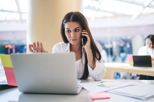 Serious female manager making smartphone call for discussing service and project during working process in office, pensive caucasian woman employee calling to support about website development