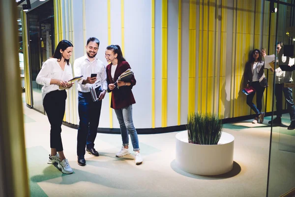Smiling adult people in casual clothes with papers in hands standing and focusing on smartphone in corridor of spacious modern office