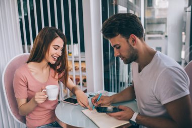 Neşeli, genç ve çekici bir kadın kahve içip defterine işaret ederken ciddi bir adam kafeteryada arkadaşça bir toplantı sırasında not alıyor ve akıllı telefon tutuyor.