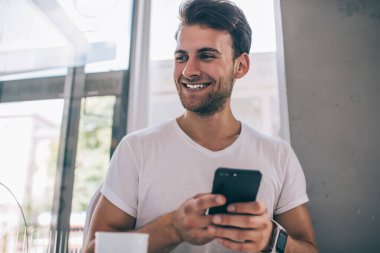 Modern kafede otururken ve eğlenirken gözlerini kaçırıp cep telefonu kullanan heyecanlı, genç ve yakışıklı bir adam.