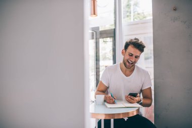 Akıllı telefon kullanan, kafede çalışırken komik mesajlara gülen ve not defterine not alan neşeli, genç bir serbest çalışan.