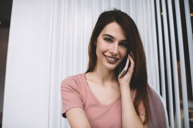 Uzun siyah saçlı, sıradan pembe tişörtlü genç bir kadın kafede oturmuş kameraya bakarken telefonla konuşuyor.