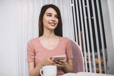 Genç, koyu renk saçlı, sıradan pembe tişörtlü, akıllı telefonlu bir kadın kafede oturmuş kahve içerken gözlerini kaçırıyor.