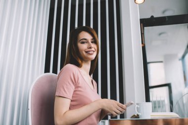 Cafe 'de boş zamanlarında kameraya bakarak akıllı telefon kullanan neşeli, genç bir bayandan.