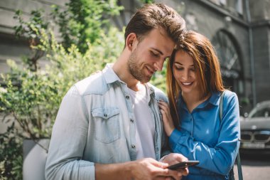 Gündelik kıyafetlerle gezen neşeli yetişkin bir adam akıllı telefonuyla etkileşime girerken çekici kız arkadaş ekranlara odaklanıyor ve caddede eski gri binaya ve güneş ışığında yeşil ağaçlara karşı dururken gülümsüyor.