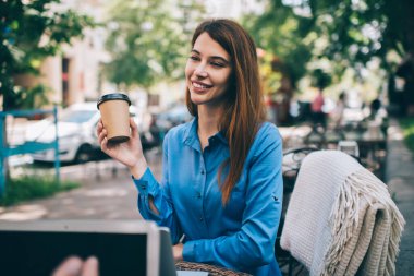 Gülümseyen, uzun saçlı, mavi elbiseli, elinde kahve fincanı olan bir kadın sıcak ekoseyle masaya oturuyor.