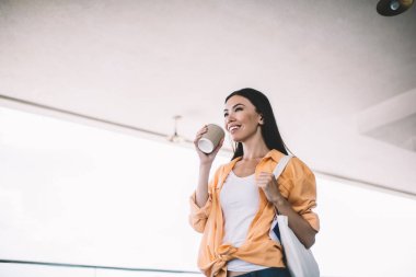 Alçak açılı, gülümseyen, modaya uygun, turuncu ve beyaz giysili, çantalı, elinde kahve fincanıyla başka yere bakan genç bir kadın.