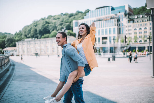 Side view of happy young man carrying girlfriend on back while spending summer holiday together and walking on square next to old building