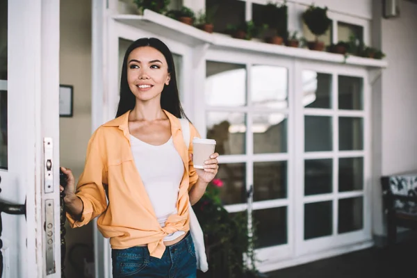 面带微笑 身穿橙色衬衫的年轻女性 白天打开咖啡店大门时 举着一杯咖啡 望着远方 — 图库照片