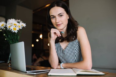 Çekici, zeki, gülümseyen kadın yazar. Elinde çenesiyle otururken kameraya bakarak notlar yaratıyor.